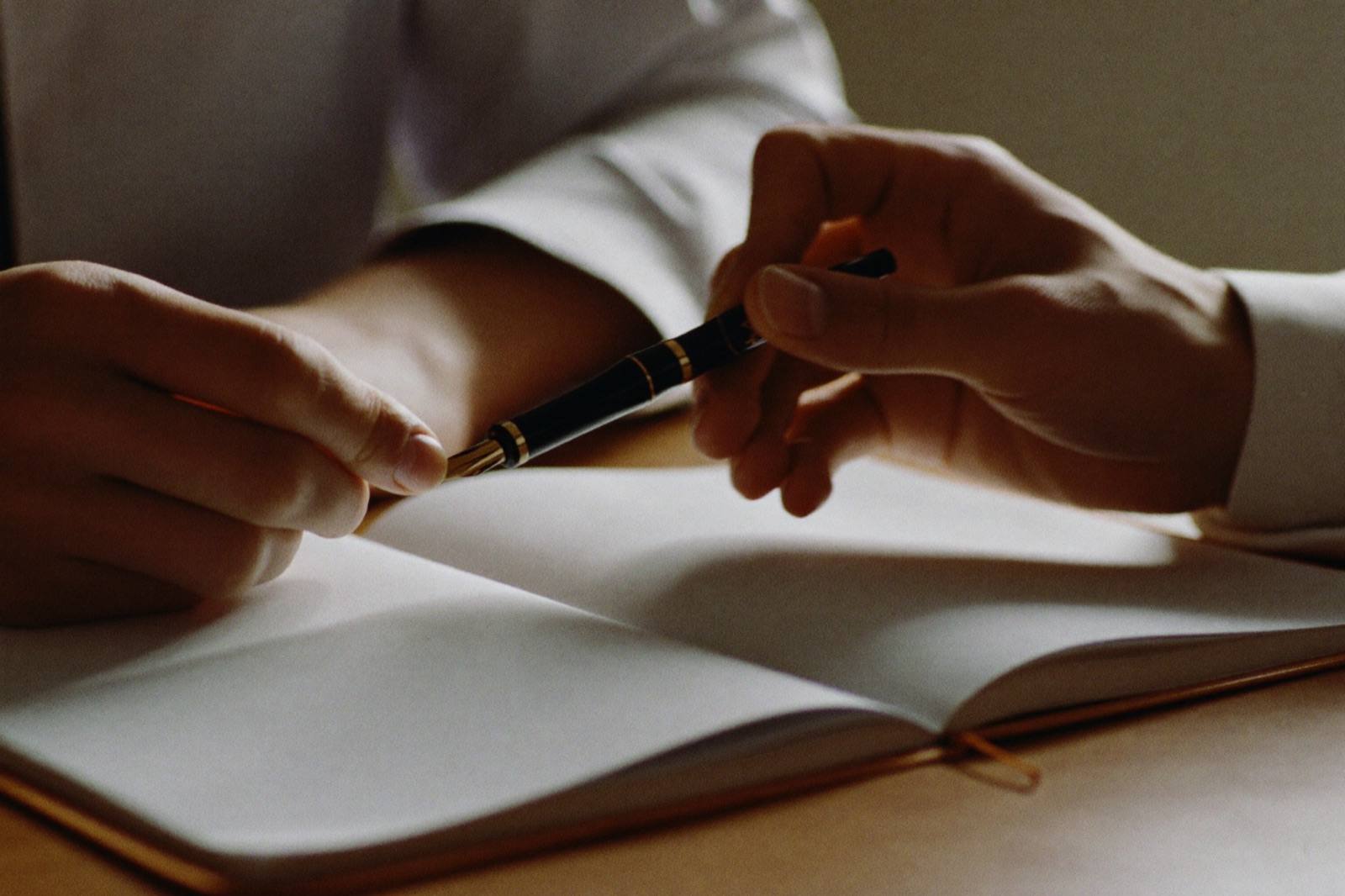 Two hands exchange a fountain pen above an open blank signing book.