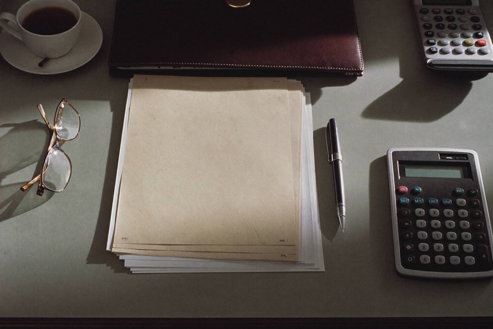 An underwriter’s desk from above: blank application papers on a leather folio, a pen, folded reading glasses, calculators and a cup of coffee, evenly spaced.